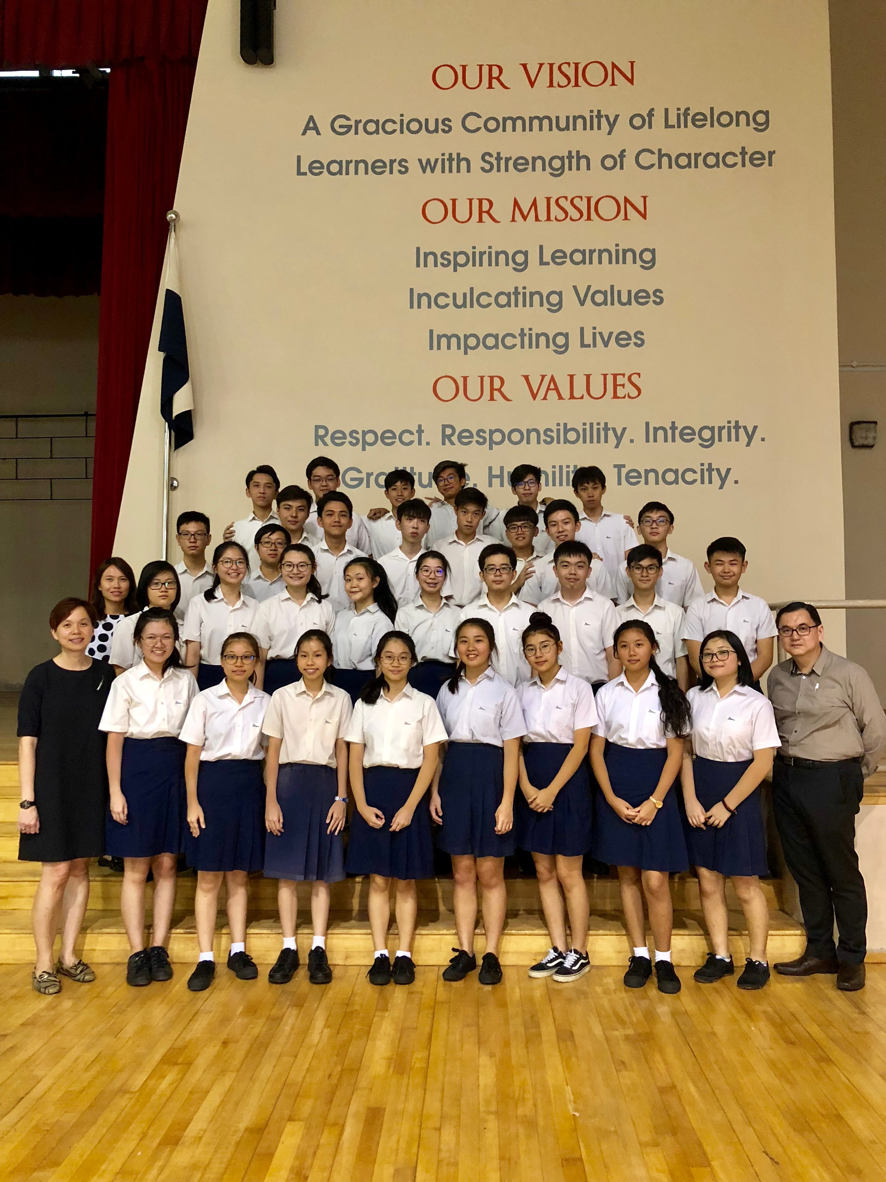 Students in white uniforms pose with two teachers in front of a school vision and mission wall display.